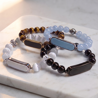 Four beaded stretch bracelets with rectangular metallic bezel settings, arranged on a white marble slab. The bracelets feature light blue, white, black, and tiger's eye beads with silver-toned and dark metallic accents.