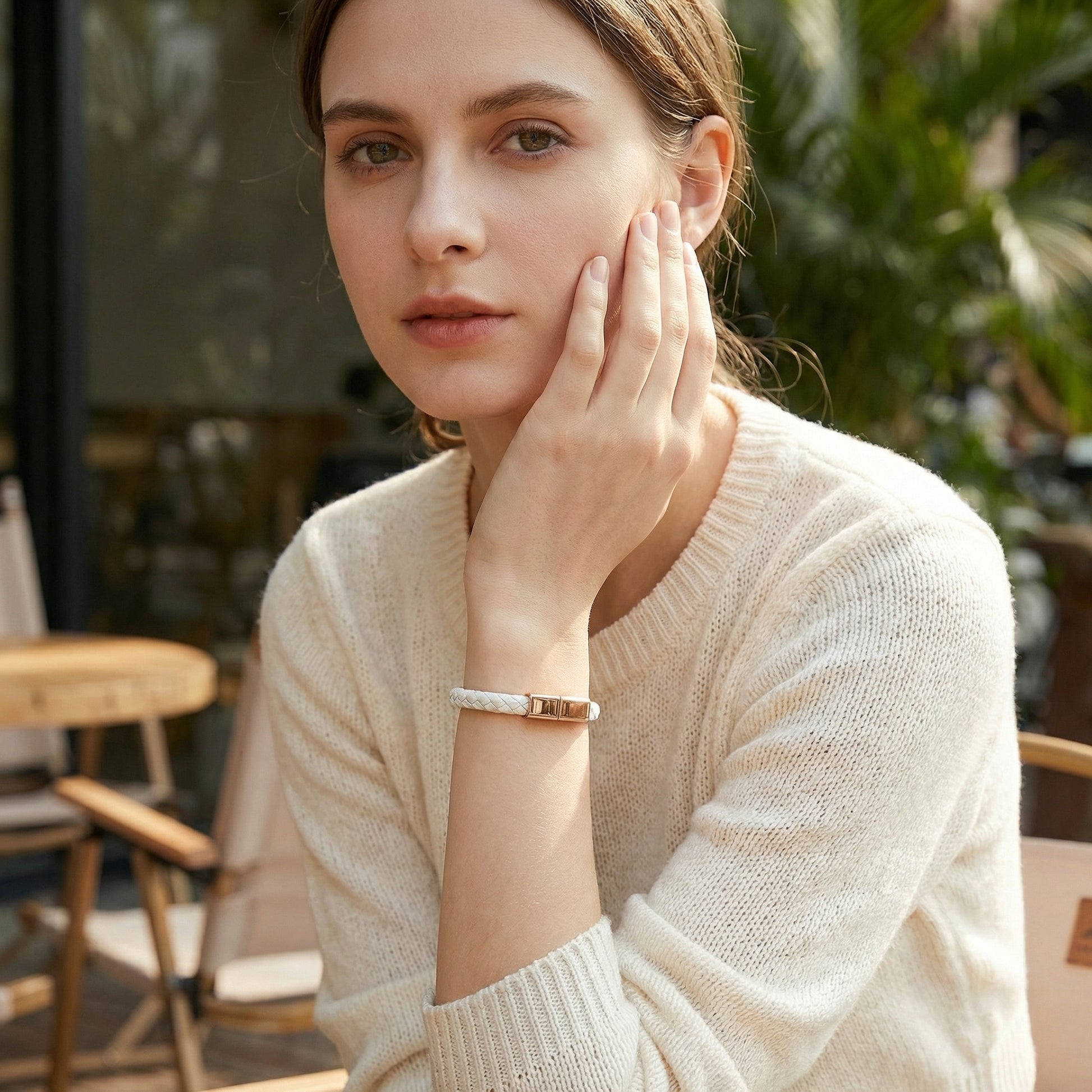 Woman in a beige sweater sitting outdoors wearing a white charging bracelet, with plants in the background.